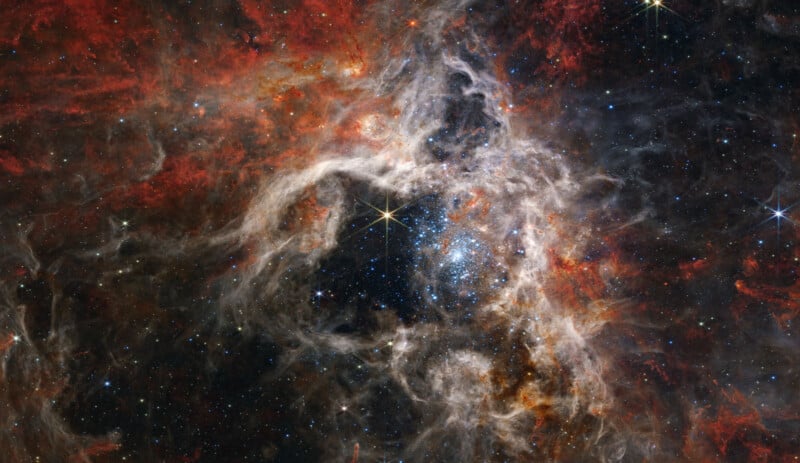 A stunning image of the Tarantula Nebula captured by the James Webb Space Telescope, showcasing a vivid, complex tapestry of bright stars, swirling gas, and dust clouds in hues of red, orange, and black, set against a dense star-studded background.