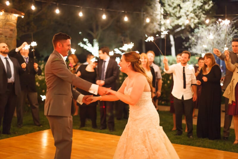 Bride and groom dance under string lights at an outdoor wedding reception. Guests stand around, holding sparklers, smiling, and watching the couple. The scene is festive, with warm lighting creating a cozy atmosphere.