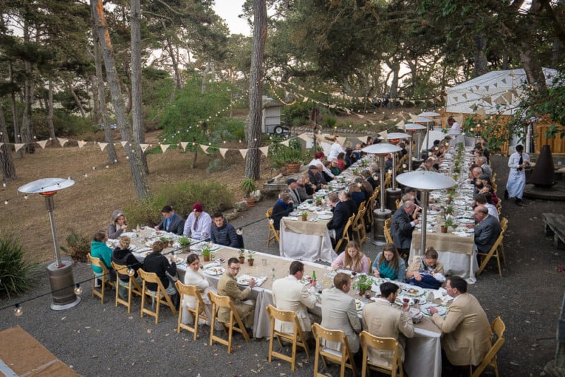 A long outdoor dinner setup with multiple tables aligning lengthwise, seated with numerous guests enjoying a meal. The scene is decorated with white string lights, rustic table settings, and surrounded by trees and greenery, creating a festive and inviting atmosphere.