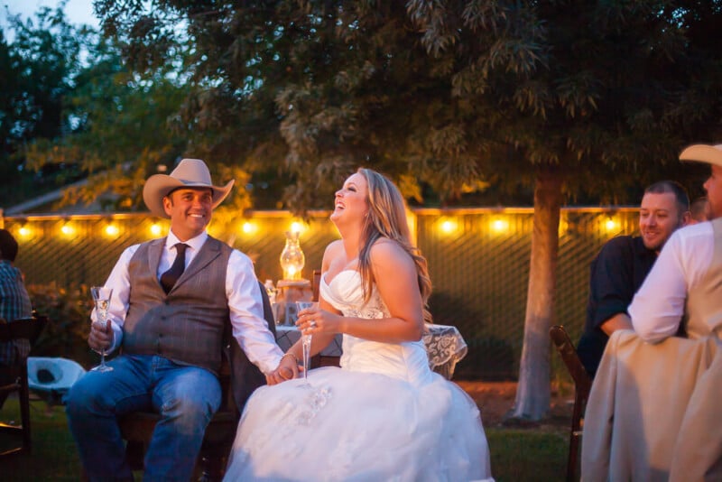 A couple sits outdoors at what appears to be a wedding reception. The woman, dressed in a white wedding gown, is laughing heartily while holding a glass of champagne. The man, dressed in a vest, tie, and cowboy hat, smiles and holds her hand. String lights are visible in the background.