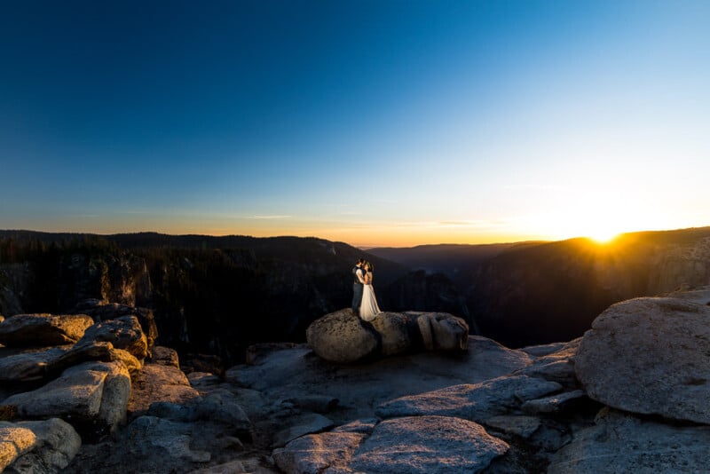 A couple in wedding attire stand on a large rock at sunset, surrounded by a rocky landscape. The sun is setting behind distant mountains, casting a golden light across the scene, creating a romantic and dramatic atmosphere.