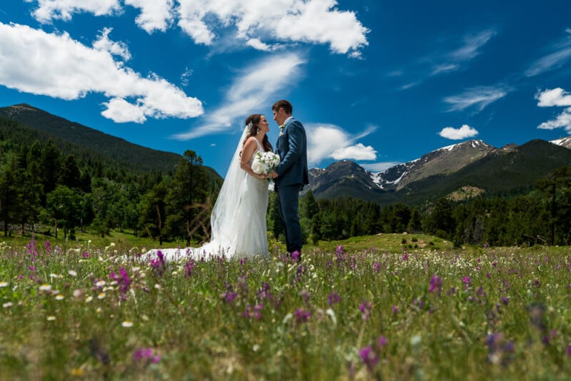 Bride and groom stand face-to-face in a meadow of wildflowers, surrounded by green hills and snow-capped mountains under a bright blue sky with scattered clouds. The bride wears a white dress and veil, holding a bouquet, while the groom is in a blue suit.