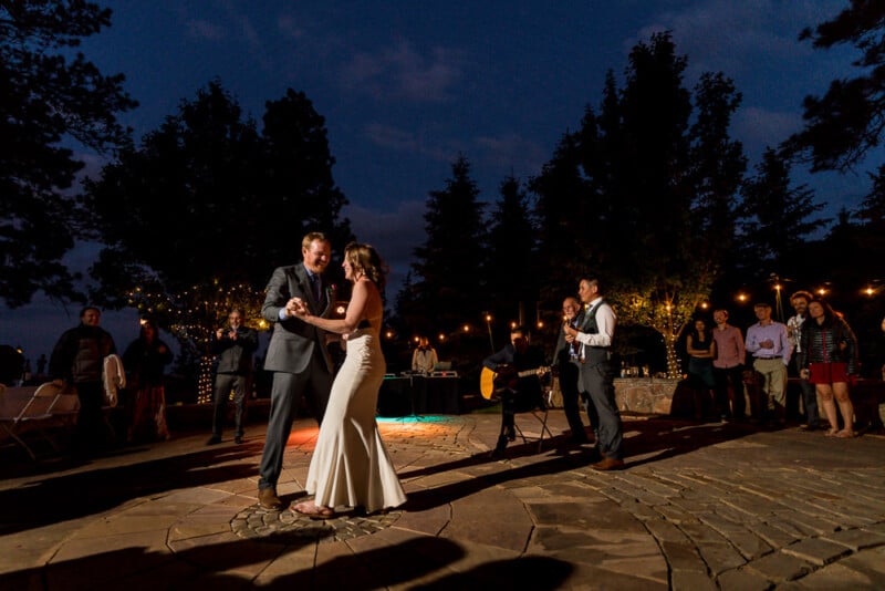 A bride and groom share their first dance at an outdoor evening wedding reception. They are illuminated by string lights and are surrounded by guests, trees, and a stone patio. Festive lights and musicians enhance the romantic ambiance.