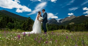 A bride and groom stand facing each other in a picturesque meadow filled with wildflowers, with a backdrop of mountains and a partly cloudy blue sky. The bride holds a bouquet and wears a long veil, while the groom is dressed in a suit.