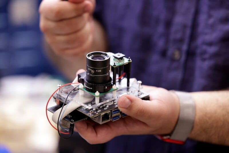 A person is holding a handmade electronic device with a camera module attached to a circuit board. Various wires are connected to the board, and the person's hand is adjusting a component on the device. They are wearing a wristband and a short-sleeved shirt.