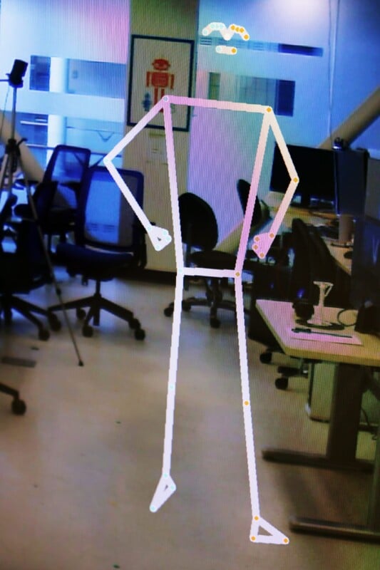 A colorful stick figure overlay is shown superimposed on the image, standing in an office space with empty desks, chairs, and office equipment in the background. The figure has distinct straight lines representing limbs, a torso, and a head outline.