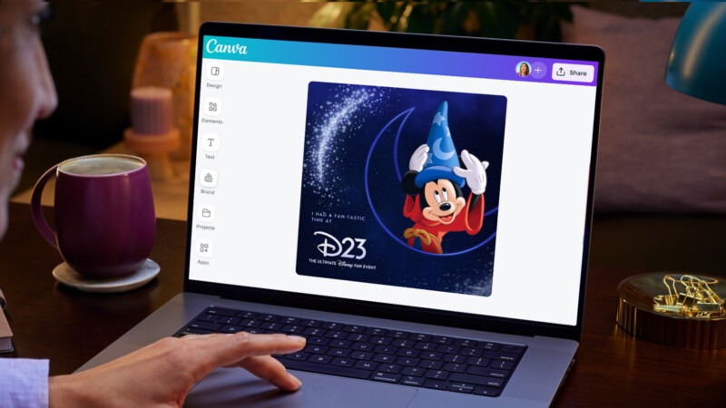 A person uses a laptop displaying Canva's design interface. On the screen is an image of Mickey Mouse dressed as a wizard with a hat, and text reading "D23: The Official Disney Fan Club." Beside the laptop, there is a pink mug, a golden lamp, and other desk items.