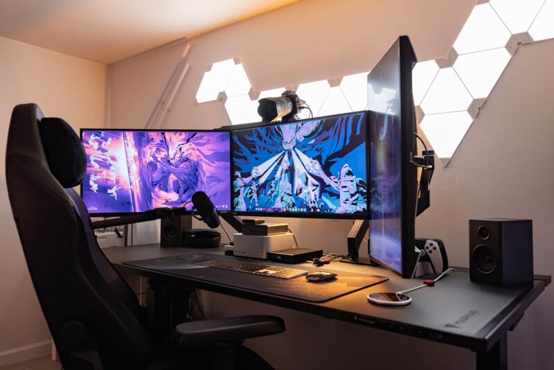 A sleek gaming and streaming setup featuring three large monitors with vibrant, abstract artwork. The desk also holds a microphone, speakers, and a compact computer. LED panels illuminate the space, creating an ambient lighting effect.