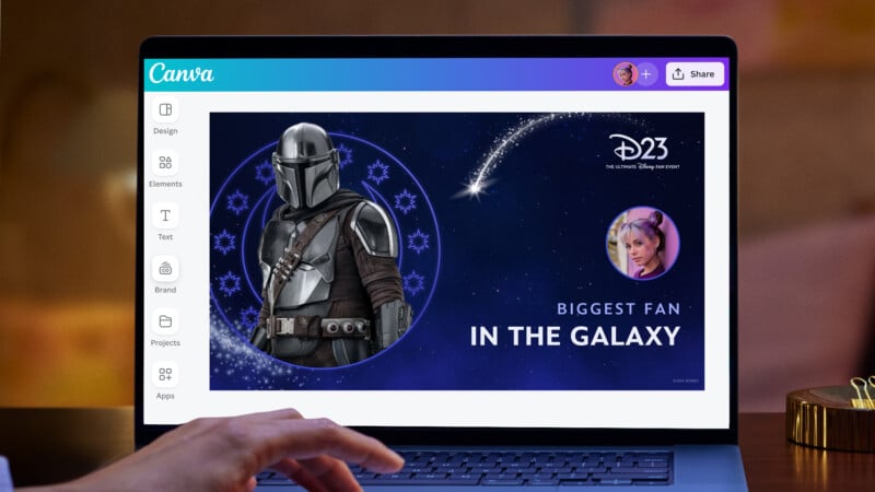 A person is working on a laptop displaying a Canva design. The design features a character in a space helmet and armor, a person with short blue hair, and text that reads "D23" and "BIGGEST FAN IN THE GALAXY" against a starry background with a comet-like graphic.