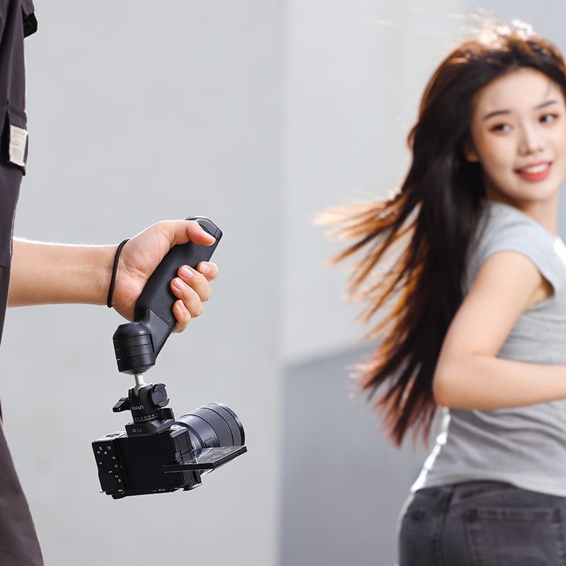 A person holds a handheld camera stabilizer with a camera attached, capturing a smiling woman with long hair walking in the background. The blurred motion effect suggests the woman is in motion. Both individuals are outdoors, possibly in an urban environment.