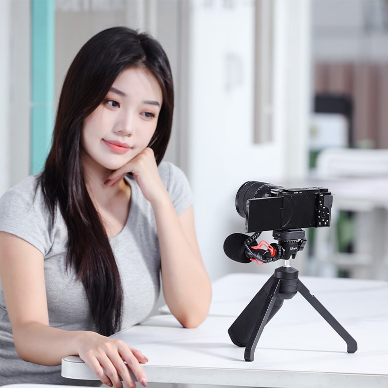 A woman with long black hair, wearing a gray short-sleeve shirt, sits at a white table and gazes at a camera set up on a small tripod in front of her. The camera has a microphone attached. The background is a light-filled indoor setting.