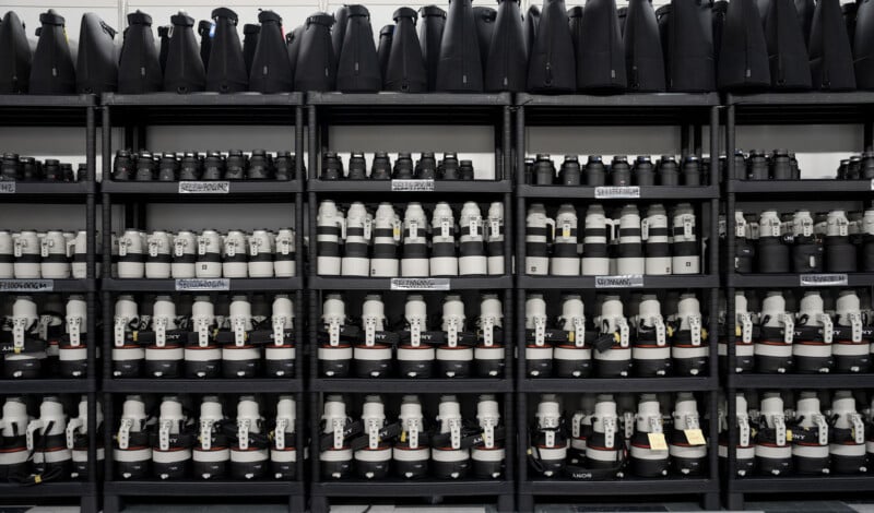 A series of black shelves holding rows of camera lenses of various sizes. The middle rows display large white telephoto lenses, while the top row has lens cases, and the bottom row features medium-sized lenses. Labels are affixed to the shelves for organization.