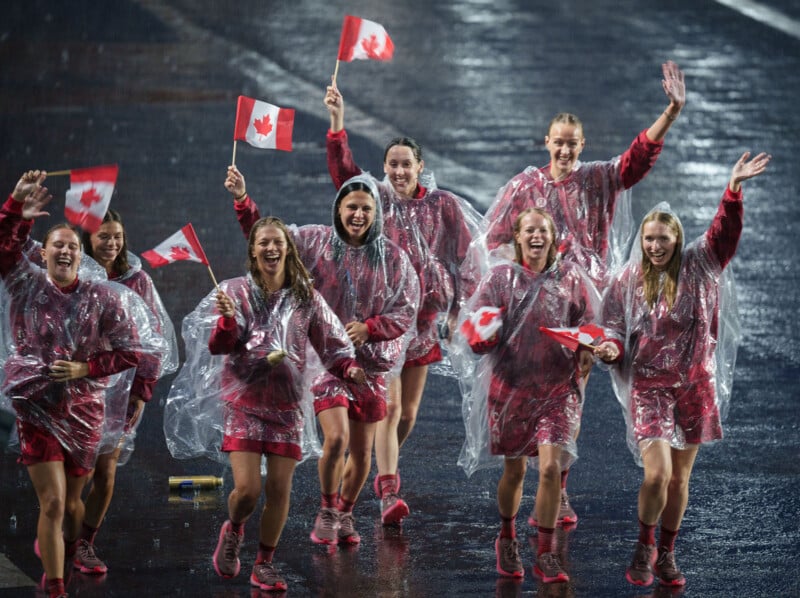 A group of women athletes in matching outfits and clear plastic ponchos enthusiastically wave Canadian flags while walking on a rain-soaked surface. They are smiling and appear celebratory, with some waving directly at the camera.