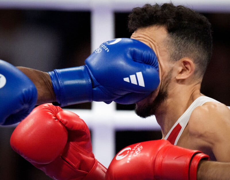 A boxer wearing red gloves gets punched in the face by an opponent wearing blue gloves during a match. The impact of the punch causes the red-gloved boxer to grimace, with the blue-gloved fist partially obscuring their face. The background is blurred.