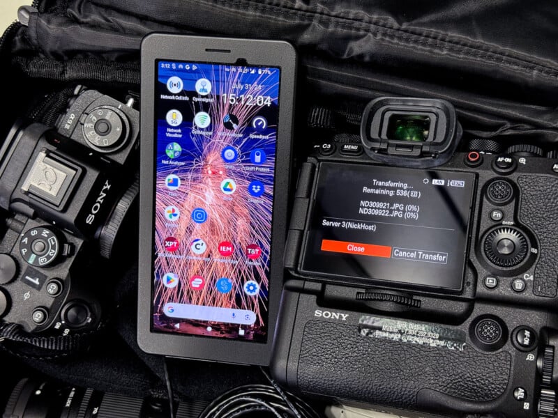 A smartphone displaying its home screen is placed between two Sony cameras inside a camera bag. The camera on the right shows a screen indicating photo transfer in progress. Various camera accessories are visible around the devices.