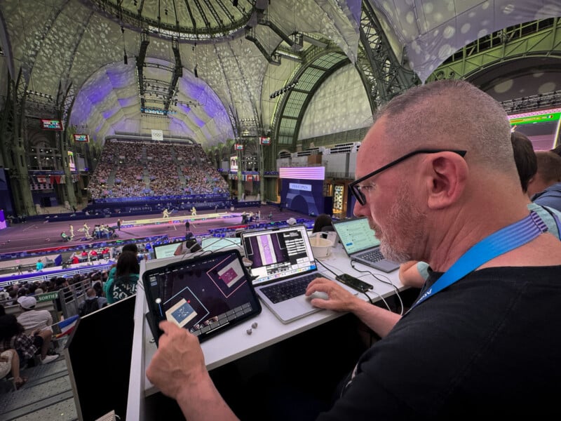 A man with glasses sits at a desk inside a large, illuminated sports arena, working on a laptop and tablet. The arena is filled with spectators, with athletes visible on the field below. Large digital screens and intricate architectural details adorn the venue.