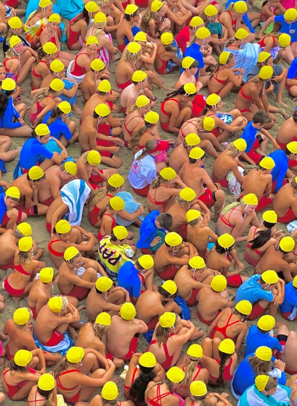 A large group of people, mostly children, sit closely together on a sandy beach. They are wearing bright yellow swim caps and red swimsuits. Some wear blue swimsuits or cover themselves with blue and white towels. The scene is vibrant and colorful.