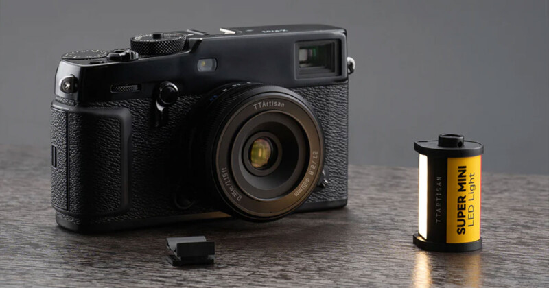 A black vintage-style camera sits on a wooden surface with a lens cap placed next to it. Beside the camera is a small roll of yellow film labeled "SUPER-MINI LCD 100R" with black and white text. The background is a plain, dark grey.