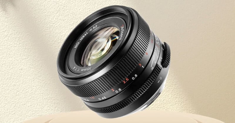 A black camera lens with a focal length of 35mm f/1.4 from 7Artisans, positioned against a light beige background. The lens features various markings for focus distance and aperture settings. The glass elements are visible, reflecting light.