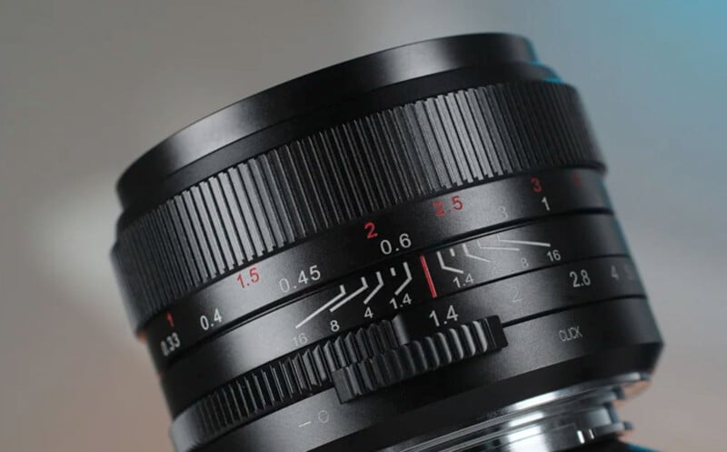 Close-up image of a black camera lens with red and white markings indicating various focal lengths and aperture settings. The lens features adjustment knobs with a textured grip pattern. The background is out of focus, highlighting the details on the lens.