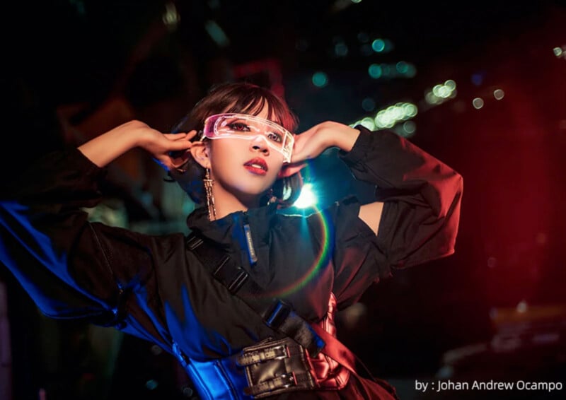 A person poses dramatically with hands placed behind their neck, wearing futuristic luminous glasses, a black outfit, and a harness. The background is filled with colorful lights creating a vibrant and dynamic atmosphere. Photo credit to Johan Andrew Ocampo.