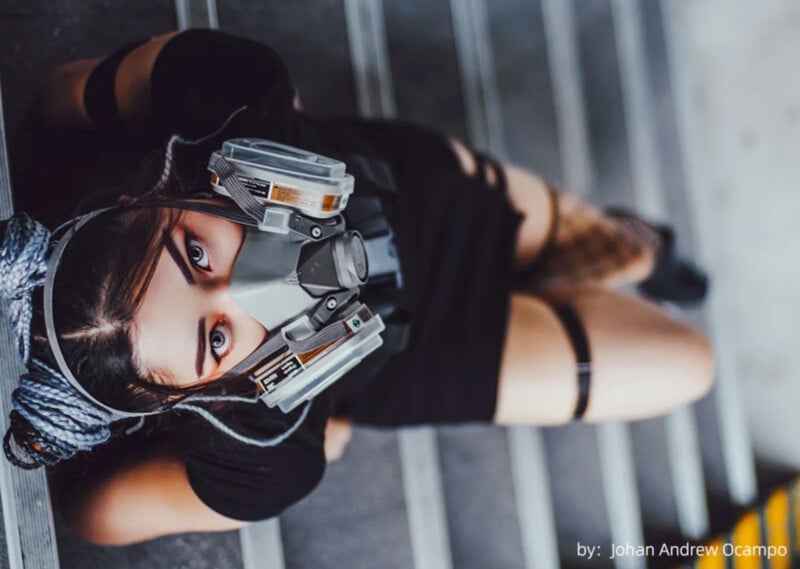 A woman with long braided hair is sitting on stairs, wearing a detailed sci-fi mask. She is dressed in black, with part of her outfit including torn fabric. The photograph, taken from above, highlights the mask and her intense gaze. Photo credit: Johan Andrew Ocampo.