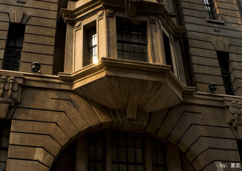 The image shows the detailed architectural facade of a historic building with stone cladding and intricate window designs. Sunlight highlights the structure, particularly around a bay window, casting shadows that accentuate the ornate stonework.