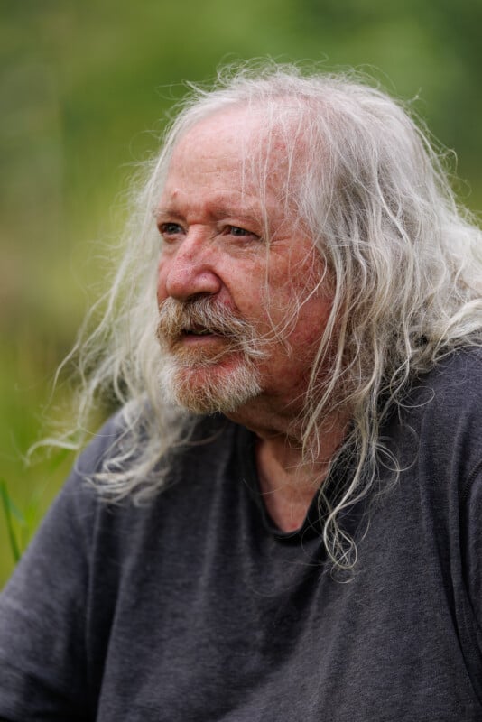 An elderly person with long gray hair and a beard is pictured outdoors. They are wearing a dark gray shirt and surrounded by greenery. The person has a thoughtful expression and the background appears to be a natural setting.