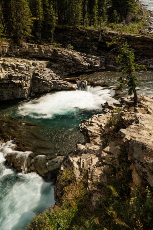 A powerful waterfall flows through rocky terrain surrounded by lush forest. The churning white water contrasts with the calm, clear blue pool below. The scene is framed by tall cliffs and scattered evergreens under a bright sky.