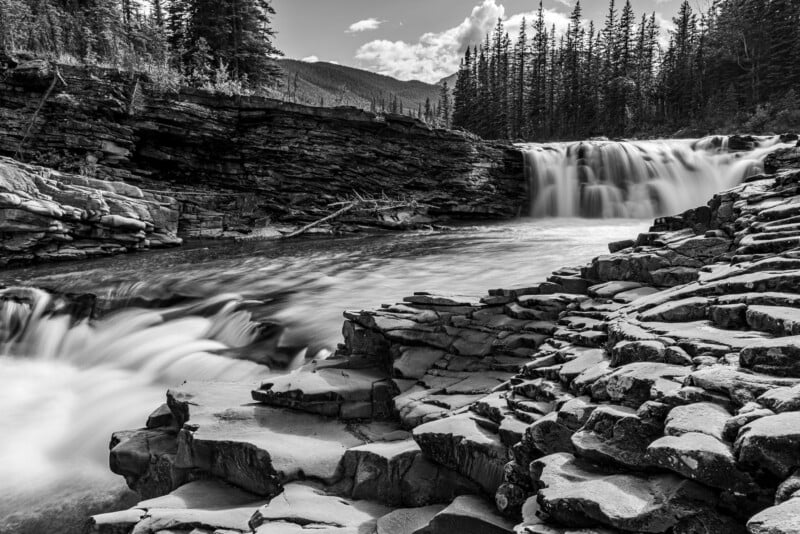 A serene black and white image of a waterfall cascading over rocky tiers into a flowing river, framed by rugged rock formations and a backdrop of tall evergreen trees and distant mountains under a partly cloudy sky.