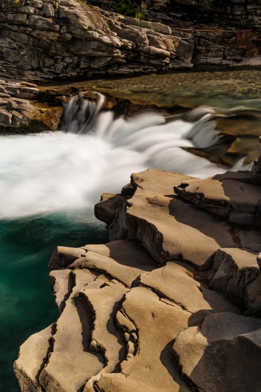 A serene waterfall cascades over smooth, layered rock formations into a clear turquoise pool. The surrounding rock faces exhibit detailed textures and natural patterns, reflecting the serene beauty and power of nature.