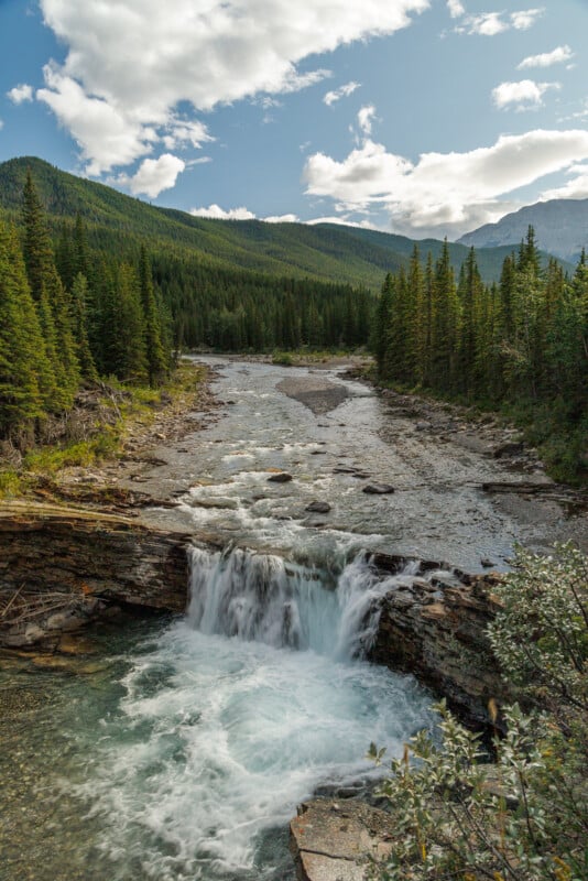 A picturesque scene with a small waterfall cascading into a clear pool of water, set against a backdrop of lush, green forest and rolling mountains under a partly cloudy sky. The calm river above contrasts with the dynamic energy of the waterfall below.