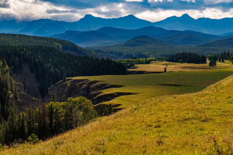 A scenic landscape featuring a grassy, rolling hillside in the foreground, dense forests below, and a series of mountain ranges stretching into the background. The sky is partially cloudy, casting dramatic shadows over the terrain.