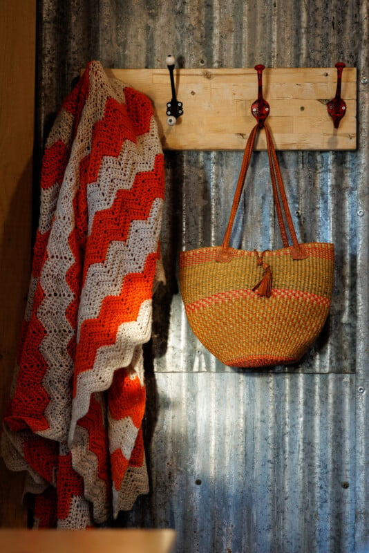 A cozy crochet blanket with red, orange, and white zigzag patterns hangs on a wooden coat rack beside a woven straw handbag with leather straps. The items are set against a corrugated metal wall backdrop, creating a warm and rustic atmosphere.