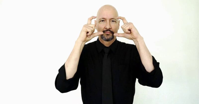 A bald man with a goatee, dressed in a black shirt and black tie, stands against a white background. He is making a hand gesture where both hands are raised, with index fingers extended and thumbs pointing upwards, creating a rectangular shape.