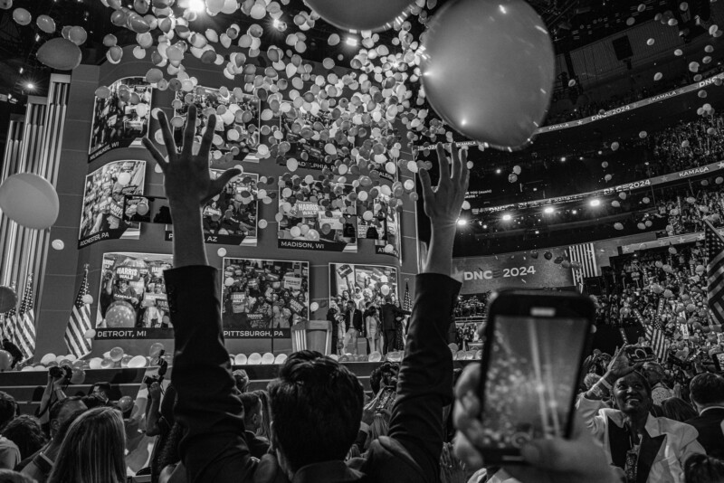 Black and white photo of a crowd celebrating at a political convention. A person in the foreground raises their hands as balloons drop from the ceiling. People take photos with their phones. Large screens display various city names like Detroit, Madison, and Pittsburgh.