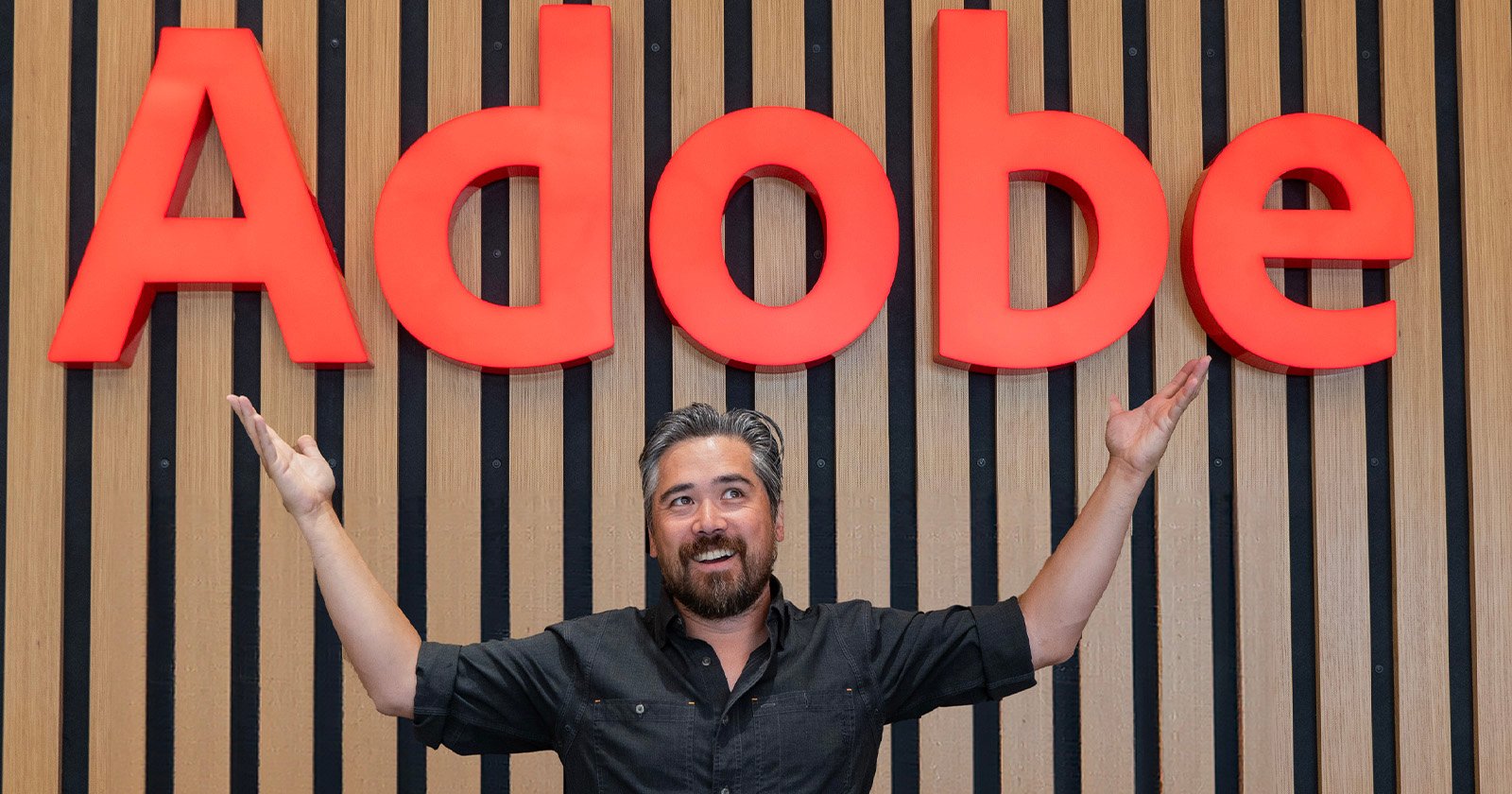 A person with a beard and dark hair stands in front of a wall with a large red "Adobe" sign, raising their arms in excitement. The background features vertical wooden slats.