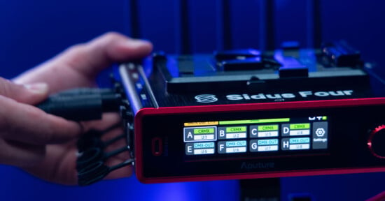 Close-up of a person’s hand plugging a cable into an Aputure Sidus Four device. The device displays a screen with various menu options and settings, and a large red dial is visible on the right side. The background is out of focus and lit with blue light.