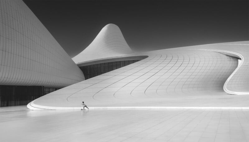 A person walks alone on a vast, empty plaza next to a large, modern architectural structure with smooth, flowing, curved surfaces and a white facade, set against a dramatic, dark sky, showcasing a stark contrast between the built environment and surroundings.