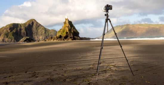 A camera mounted on a tripod stands on a deserted sandy beach with rocky hills and the ocean in the background under a partly cloudy sky.