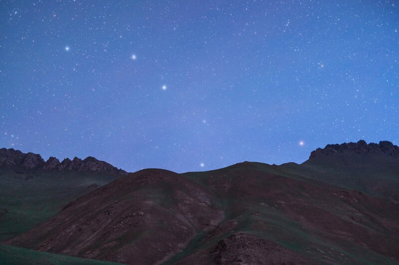 A landscape image featuring a range of smooth, rolling hills under a clear, star-filled night sky. The horizon is framed by jagged mountain peaks, and the scene is illuminated by the soft, blue light of the twilight stars.