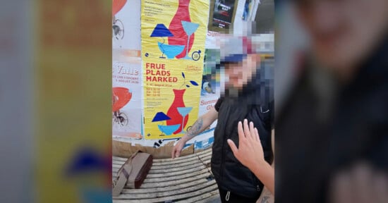 A person in a black vest and cap stands in front of colorful posters, one of which reads "FRUE PLADS MARKED." A hand, partially visible in the foreground, seems to reach towards the person. There is a suitcase or bag on a bench nearby.