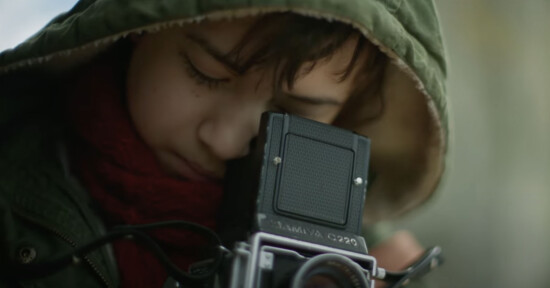 A young child, bundled in a green hooded jacket and red scarf, looks through the viewfinder of an old-fashioned camera. The child appears focused, deeply in concentration, as they prepare to take a photograph. The background is blurred, highlighting the child's intent.