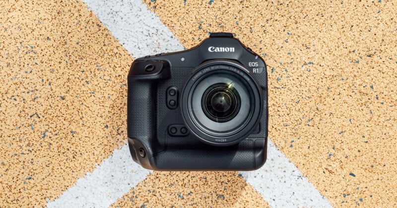 Top view of a Canon EOS R1 camera with a lens, placed on a textured, speckled background with white diagonal lines. The camera is positioned centrally, showcasing its buttons, grip, and lens details.