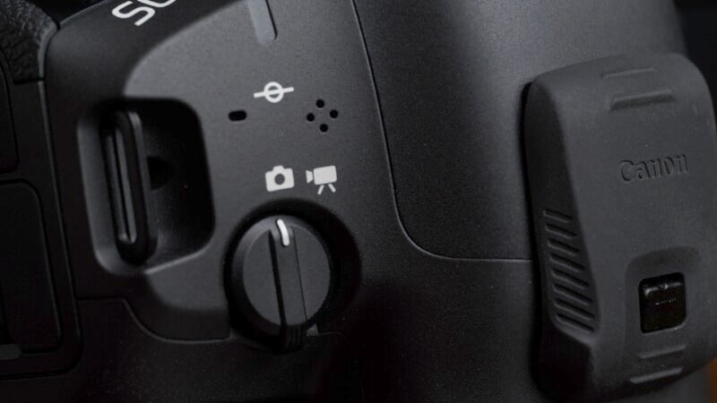 Close-up view of a camera's mode dial displaying icons for photo, video, and auto settings. The camera body is black, with the Canon logo partially visible on the right side. Various buttons and ports are also visible along the left side.