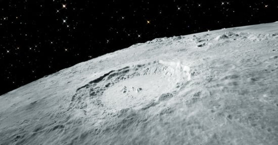 A detailed view of the moon’s surface shows a large, prominent crater surrounded by rough, uneven terrain. The dark, star-filled expanse of space forms the backdrop, highlighting the crater's stark, shadowed features and the rugged lunar landscape.