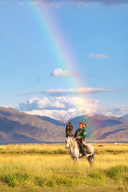 A person dressed in traditional attire sits on a white horse in a vast grassy landscape with mountains in the background. They hold a perched eagle, and a vivid rainbow arches across the sky above them.