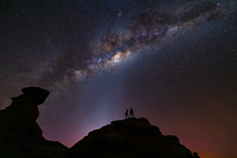 Two silhouetted figures stand on a rocky outcrop beneath a star-filled sky, with the Milky Way galaxy prominently displayed overhead. The vastness of the universe stretches out beyond them, illuminating the night with countless stars and cosmic light.