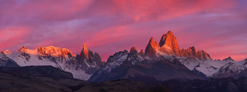 A stunning mountain range is illuminated by a vibrant sunset, casting pink and purple hues across the sky. Snow-capped peaks are bathed in a warm glow, contrasting with the dark, rocky foreground. The scene conveys a serene and majestic landscape.