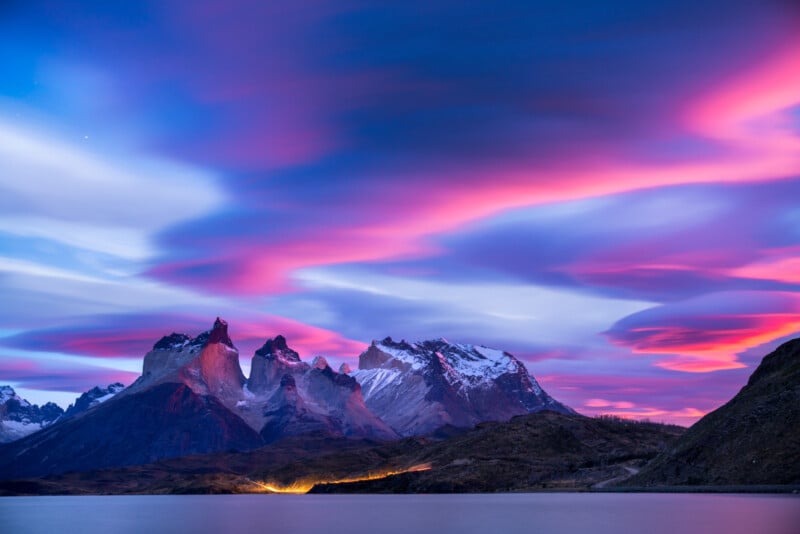 A breathtaking sunset at Torres del Paine National Park in Chile, with vivid pink, purple, and blue clouds painting the sky. Snow-capped mountains and rugged peaks are reflected in a serene lake below, creating a stunning and tranquil landscape.